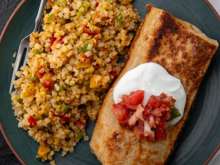 cheesy rotisserie chicken burrito on a plate with Mexican cauliflower rice garnished with Greek yogurt and salsa roja