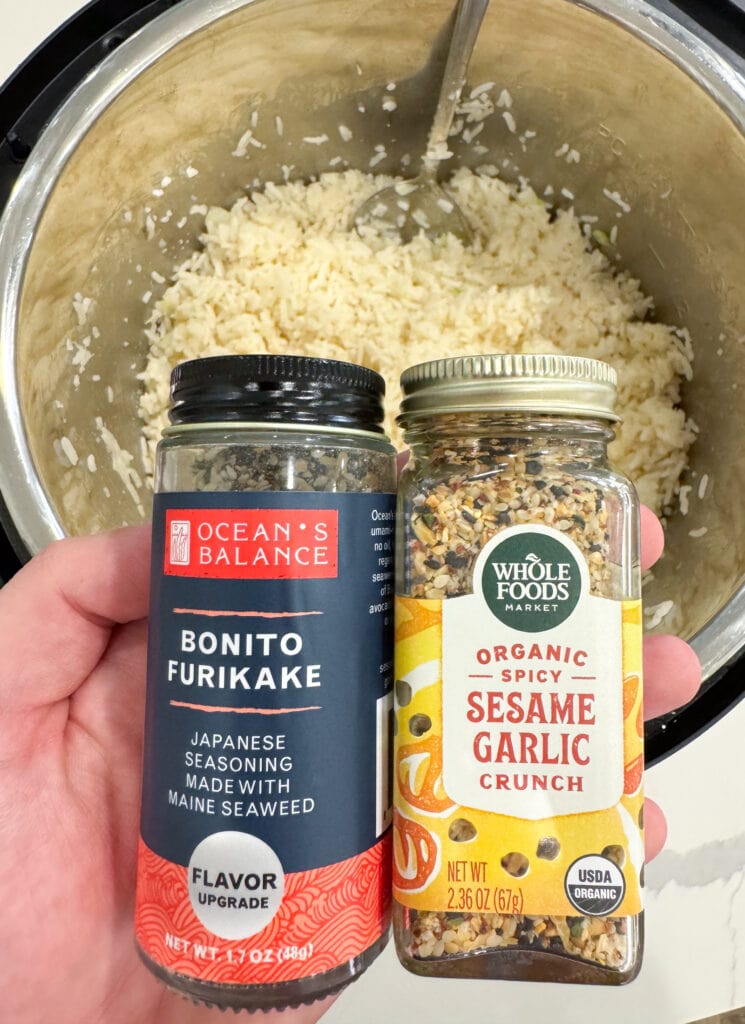 holding bottles of furikake rice seasoning and Whole Foods sesame garlic crunch above half calorie rice in an Instant Pot