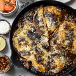 chicken mole enchilada casserole in a cast iron skillet with garnishes of cotija cheese, salsa macha, escabeche, pickled onions, limes, and jalape&ntilde;o sauce
