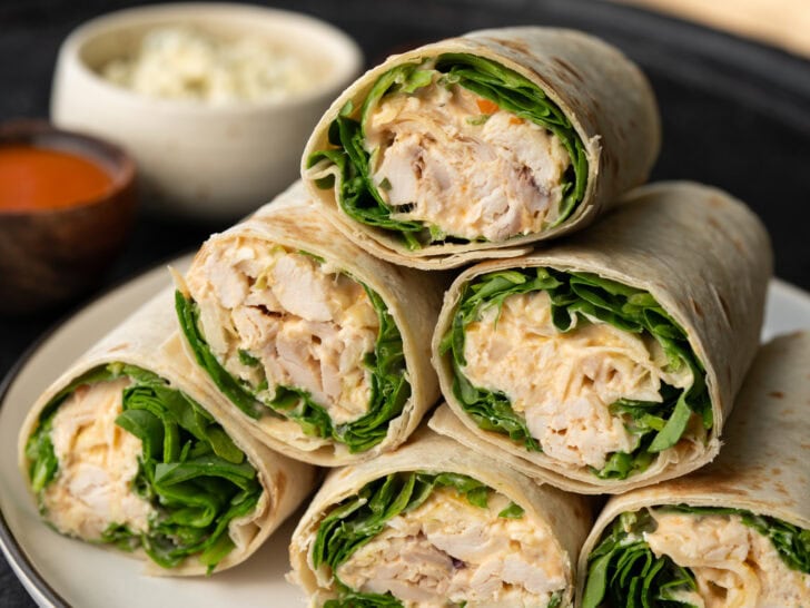 healthy buffalo chicken wraps stacked on a plate with buffalo sauce and blue cheese crumbles