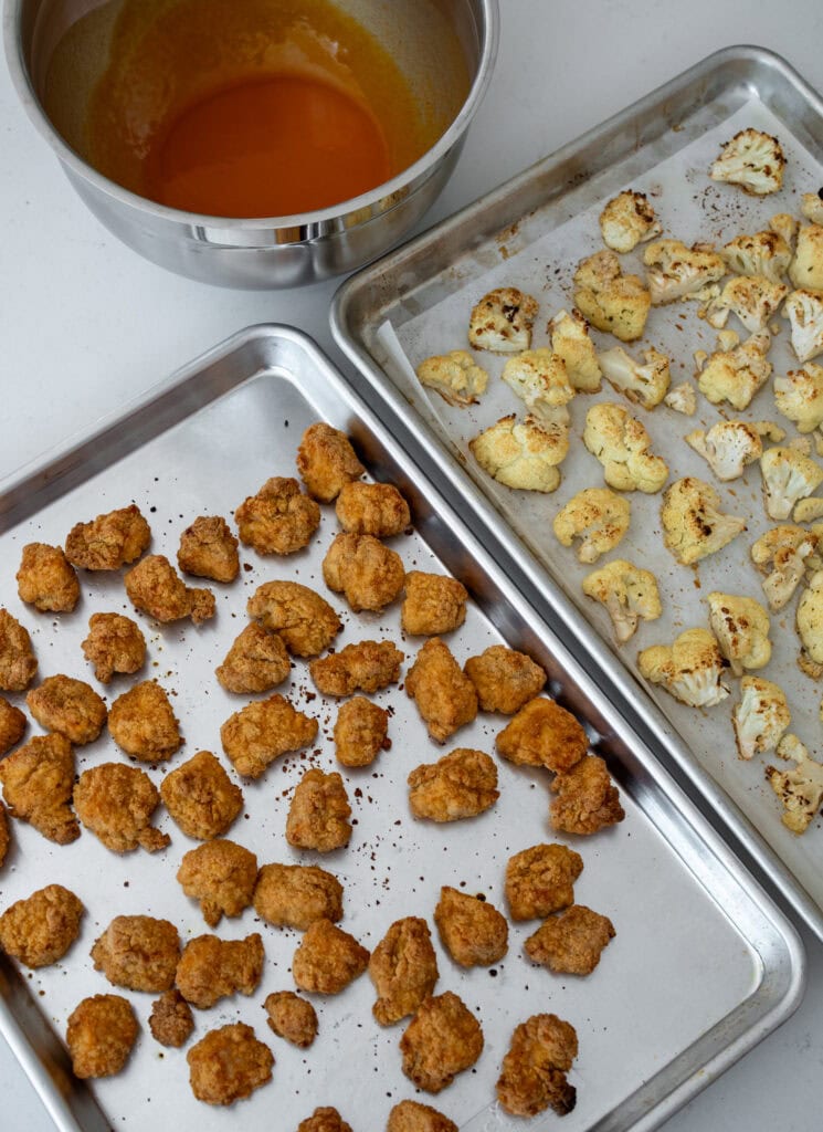 two sheet pans with baked lightly breaded chicken breast chunks and ranch roasted cauliflower florets next to a bowl with buffalo sauce