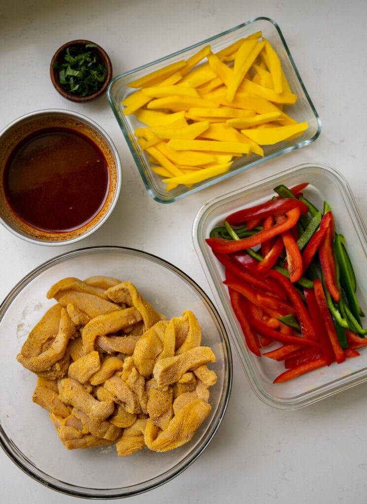 seasoned chicken breast pieces in a mixing bowl next to other bowls of Thai chili sauce, fresh Thai basil, mango slices, and bell pepper strips