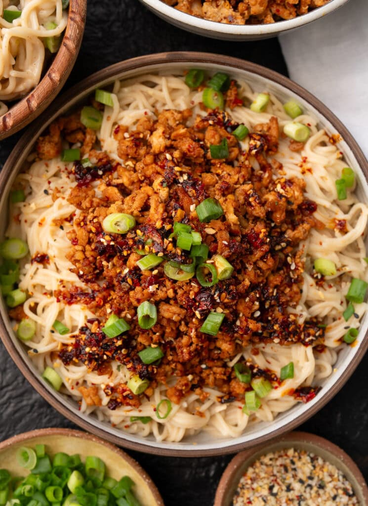 chili crisp ground chicken over peanut butter rice ramen noodles in a bowl garnished with green onions, sesame seeds, and chili oil