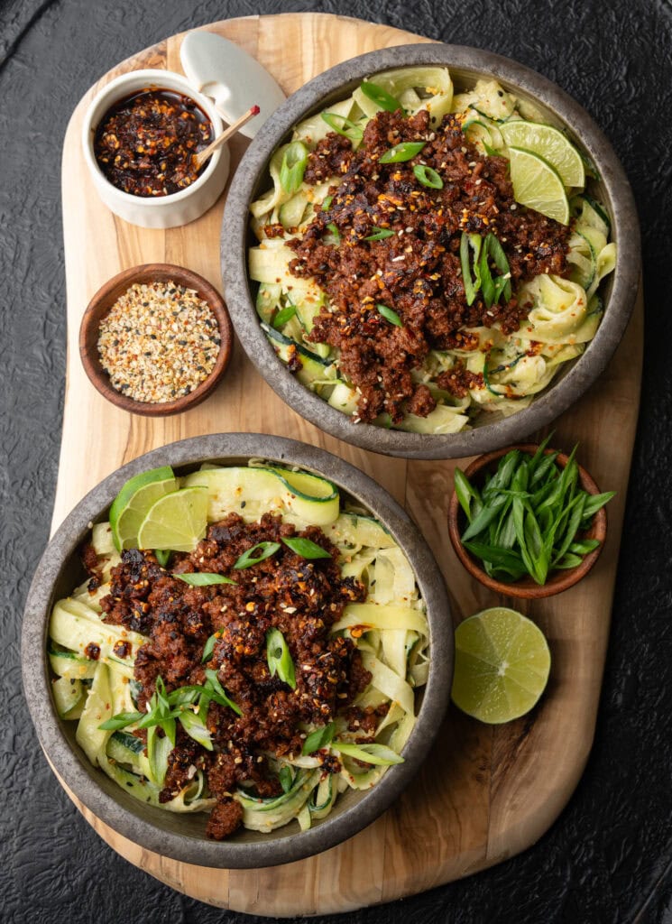 two chili crunch ground bison bowls with lime wedges, scallions, chili crunch, and sesame garlic crunch