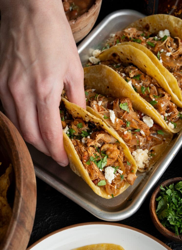 holding a chicken taco on a sheet pan with three other chicken tacos garnished with chopped cilantro and queso fresco