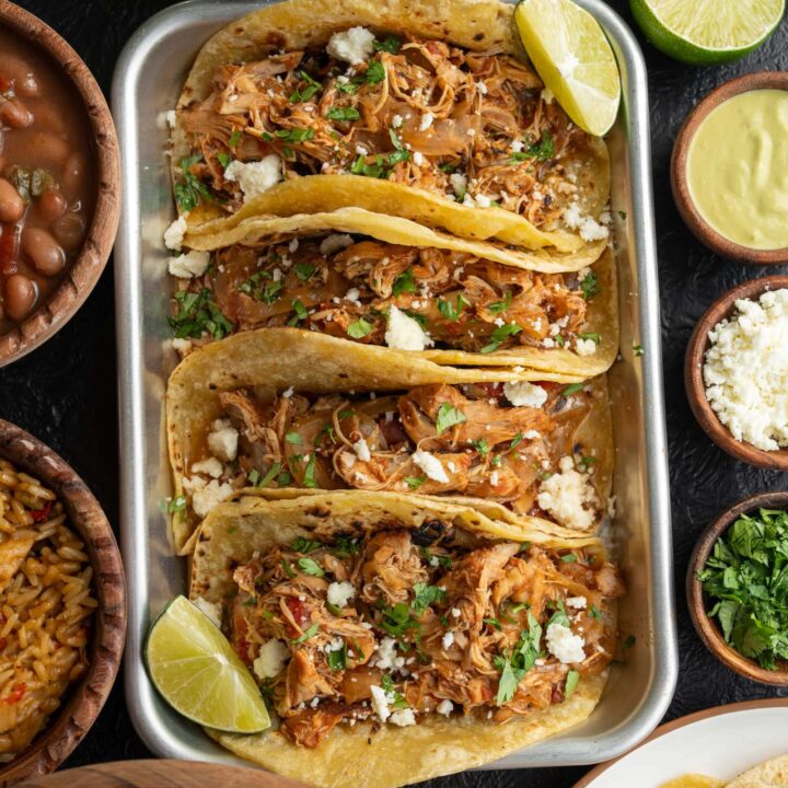 four chicken tacos on a sheet pan with chopped cilantro, queso fresco, and lime wedges