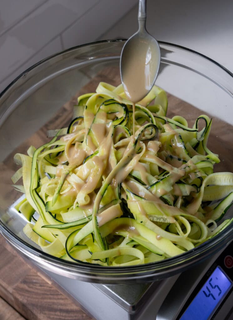 zucchini noodles in a glass mixing bowl with tahini being drizzled over the top