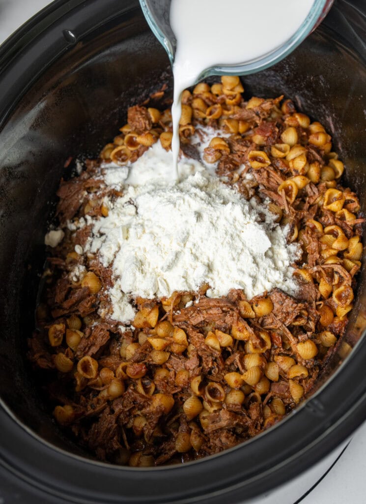 adding cheese sauce and milk to shredded beef chili mac in a Crockpot