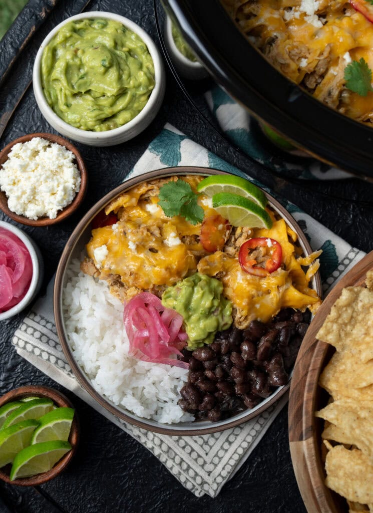 two servings of Crockpot chicken enchilada casserole in a bowl with rice, beans, guacamole, pickled onions, and lime wedges