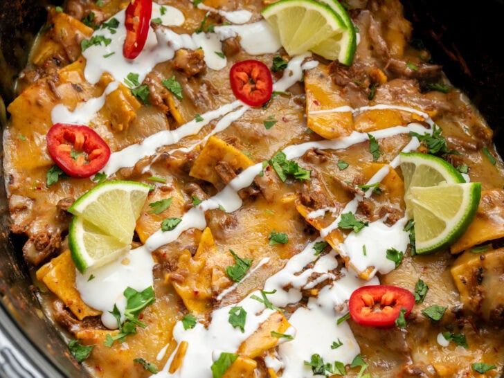 shredded beef enchilada casserole in a Crockpot garnished with Mexican crema, fresh chiles, lime wedges, and cilantro