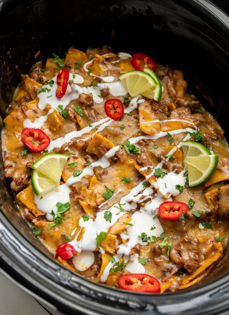 shredded beef enchilada casserole in a Crockpot garnished with Mexican crema, fresh chiles, lime wedges, and cilantro