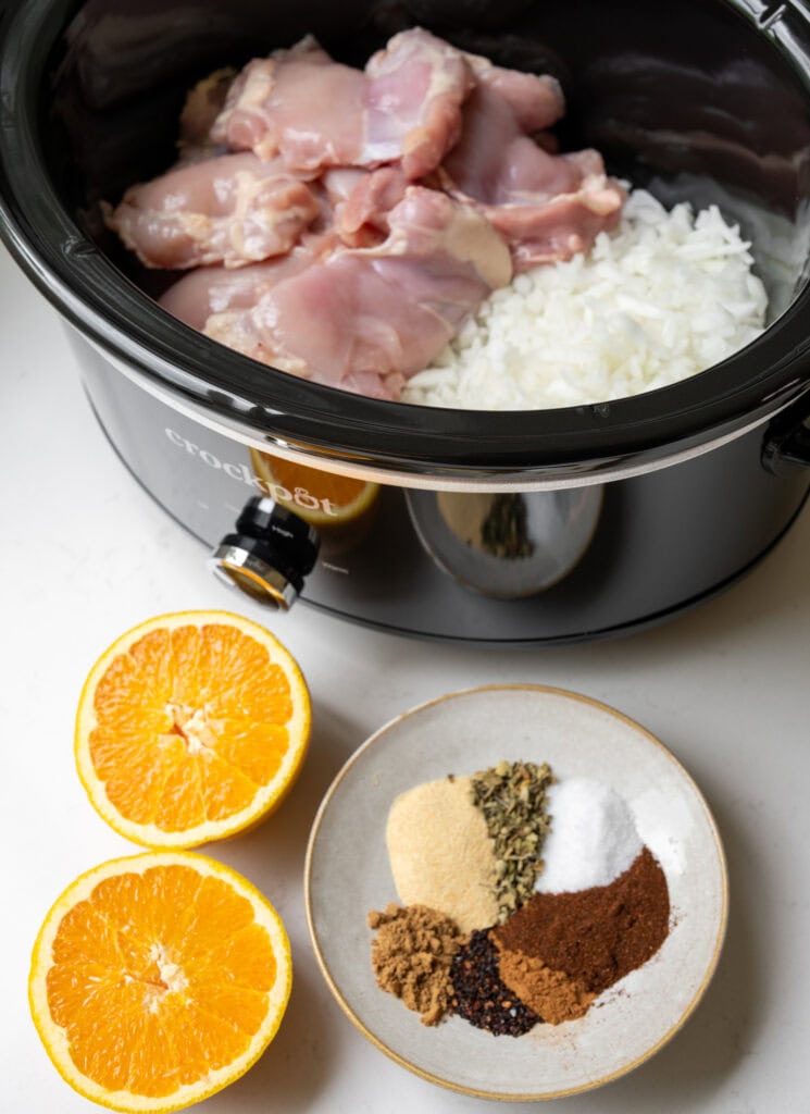 boneless skinless chicken thighs in a crockpot with diced white onion, a bowl of spices and herbs, and a halved orange