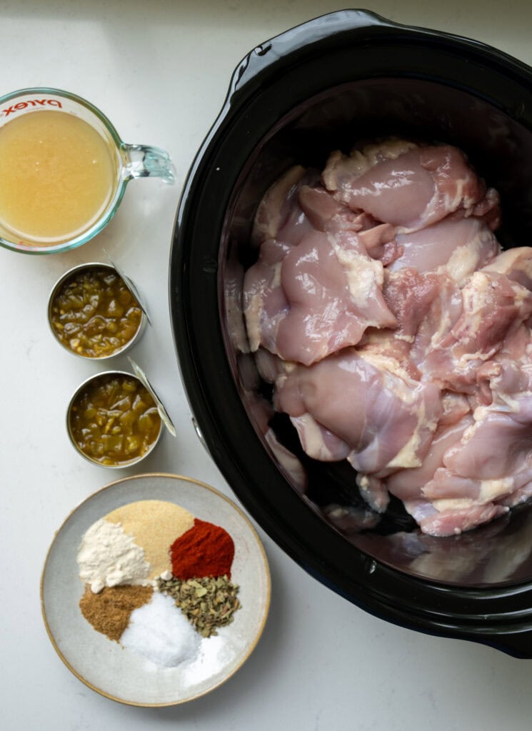 boneless skinless chicken thighs in a Crockpot beside a bowl of mixed spices, two cans of diced green chiles, and a measuring cup with chicken bone broth