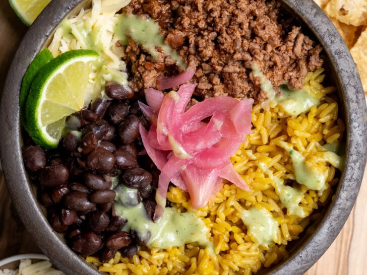 ground beef birria burrito bowl with rice, black beans, cheese, pickled onions, lime wedges, and salsa verde