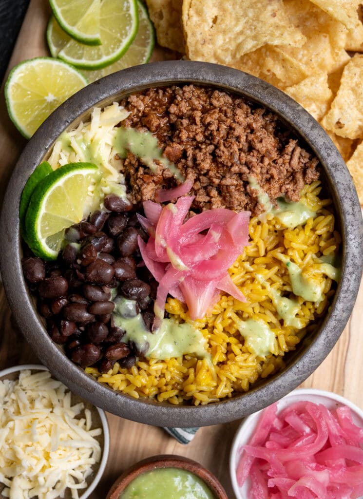 ground beef birria burrito bowl with rice, black beans, cheese, pickled onions, lime wedges, and salsa verde