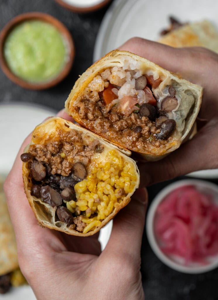 holding two halves of birria burritos with salsa verde and pickled onions in the background