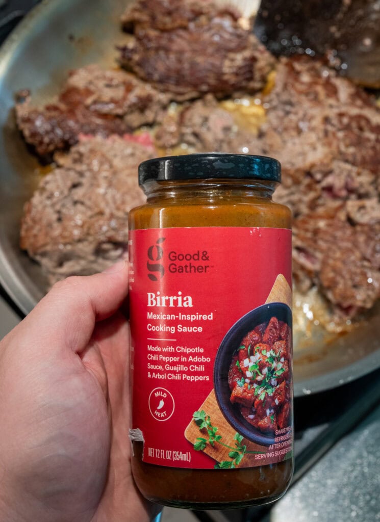 holding a jar of Good & Gather Mexican-inspired birria sauce over a skillet with browned ground beef