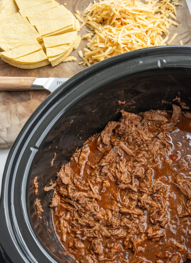 shredded top round roast in a Crockpot next to chopped corn tortillas and grated cheese