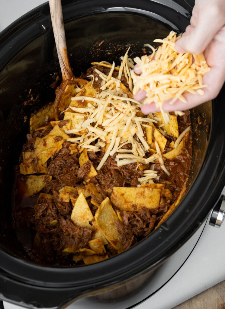 adding grated cheese on top of shredded beef and corn tortilla strips in a Crockpot