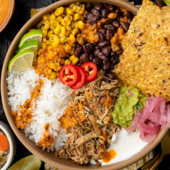 burrito bowl with Crockpot Mexican chicken, black beans, roasted corn, rice, Mexican crema, guacamole, pickled onions, lime wedges, and tortilla chips