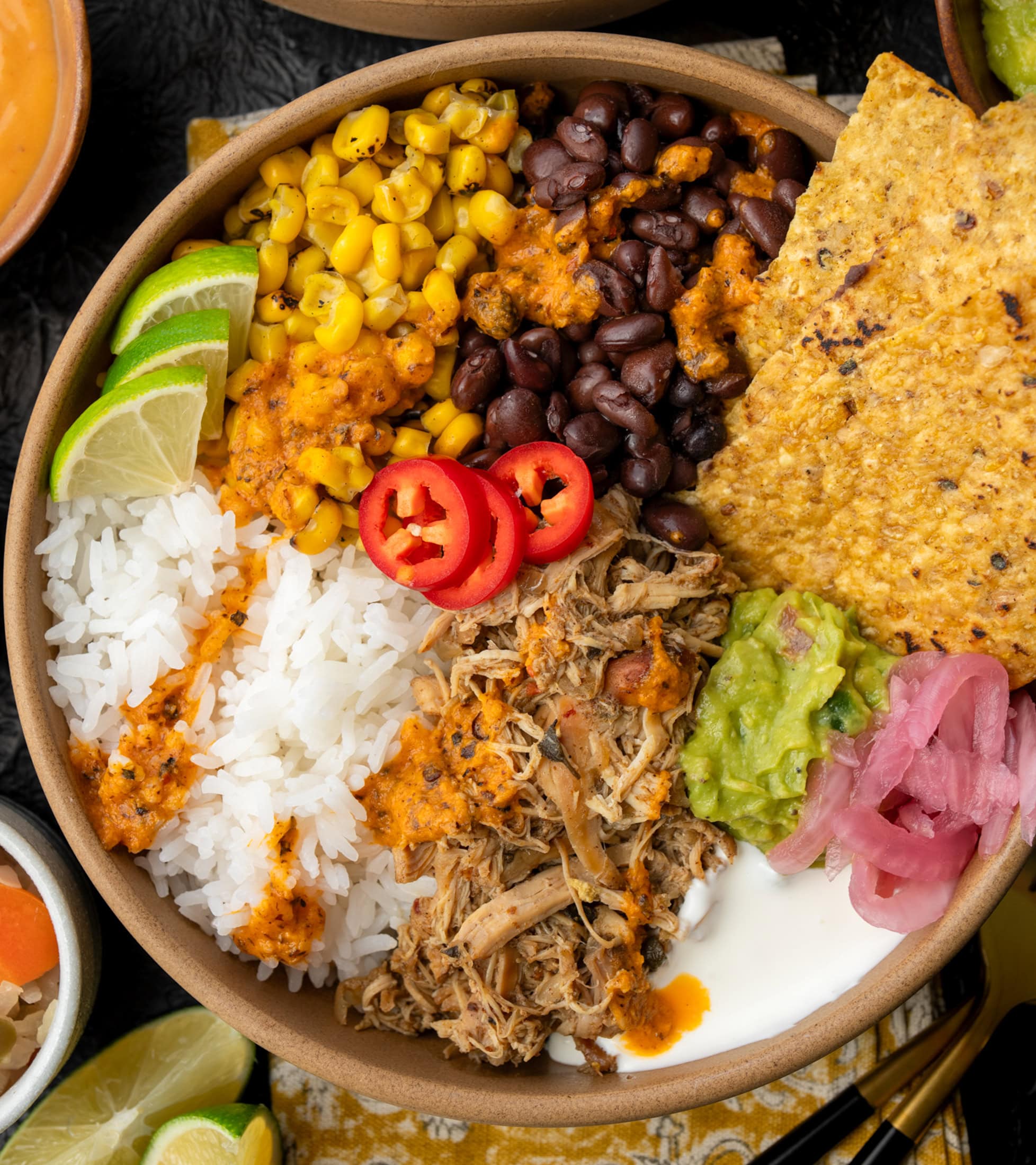 burrito bowl with Crockpot Mexican chicken, black beans, roasted corn, rice, Mexican crema, guacamole, pickled onions, lime wedges, and tortilla chips