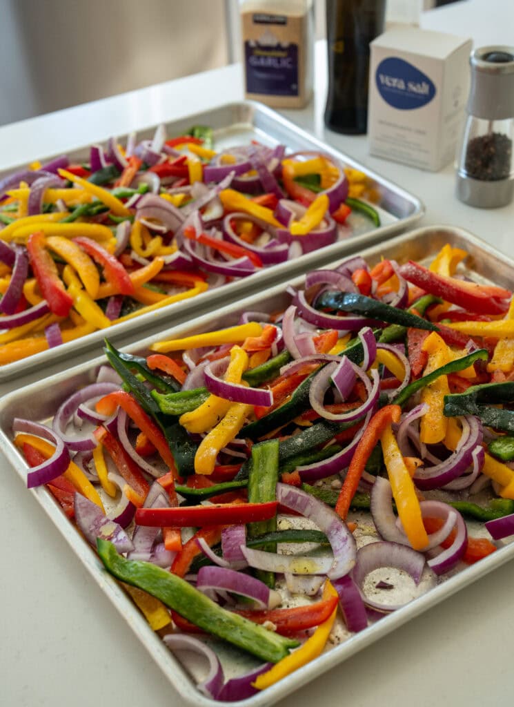 strips of peppers and onions on two half sheet pans seasoned with salt, pepper, garlic and onion powder, and olive oil