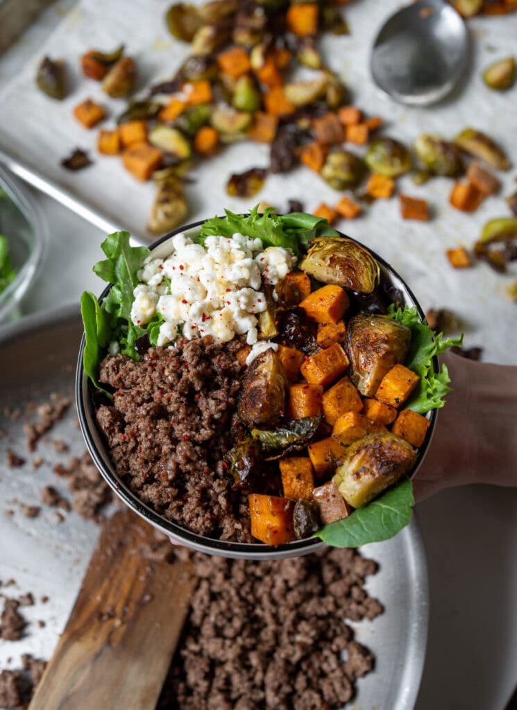 holding a bowl of garlic butter ground beef, roasted sweet potatoes and brussels, spring mix, and goat cheese drizzled with honey and chile flakes