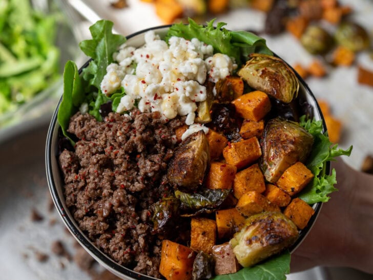 holding a bowl filled with mixed greens, garlic butter ground beef, roasted sweet potatoes and brussels sprouts, and crumbled goat cheese