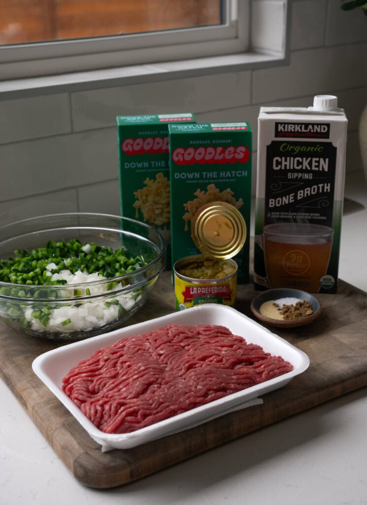 a cutting board with assembled ingredients like ground beef, diced onion and poblano pepper, Goodles down the hatch, diced green chiles in a can, spices, and chicken bone broth