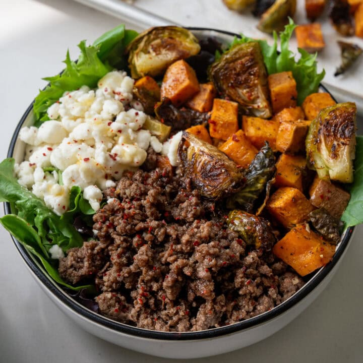 white bowl with garlic butter ground beef, roasted sweet potatoes, roasted brussels sprouts, goat cheese, spring mix, honey, and chile flakes