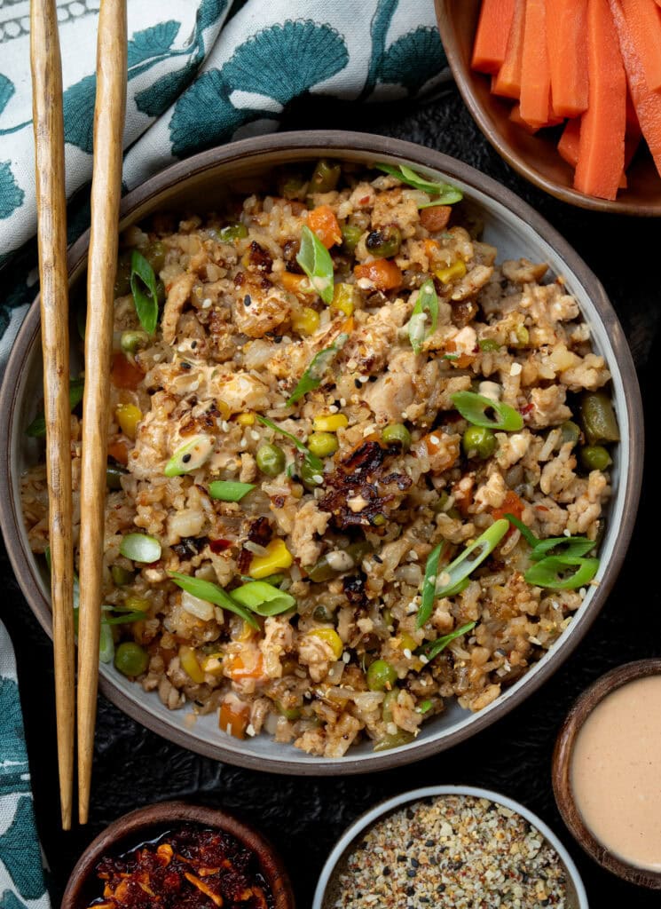 chopsticks on a bowl of ground chicken fried rice with chili crunch, toasted sesame seed garlic crunch, yum yum sauce, scallions, and pickled carrots