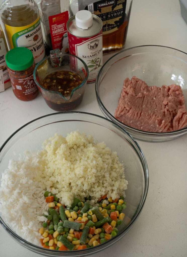 fried rice sauce ingredients next to ground chicken in a glass bowl and another glass mixing bowl with frozen rice, cauliflower rice, and mixed vegetables