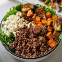 white bowl with garlic butter ground beef, roasted sweet potatoes, roasted brussels sprouts, goat cheese, spring mix, honey, and chile flakes
