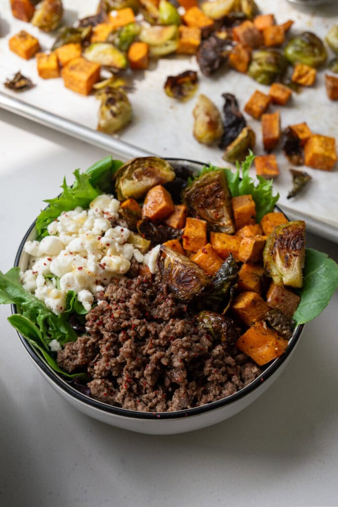 a white bowl filled with baby spring mix, goat cheese crumbles, ground beef, and roasted brussels sprouts and sweet potatoes