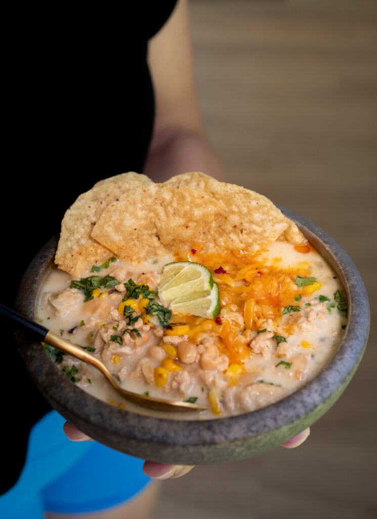 holding a bowl of white chicken chili garnished with cilantro, shredded cheddar cheese, limes, and tortilla chips