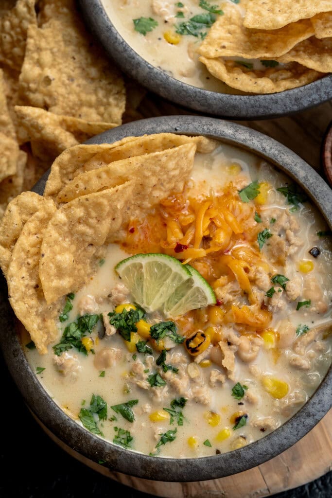 bowl of ground chicken chili garnished with tortilla chips, shredded cheddar, lime wedges, and cilantro