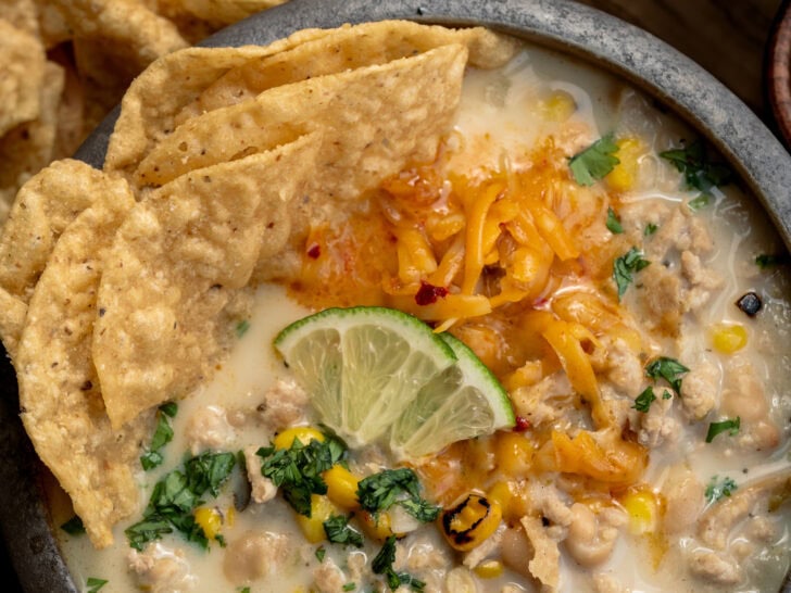 bowl of ground chicken chili garnished with tortilla chips, shredded cheddar, lime wedges, and cilantro