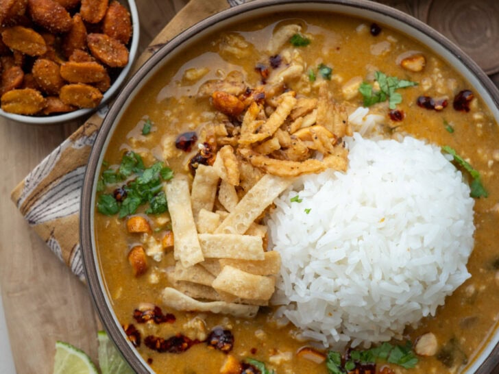 a bowl of Thai coconut ground chicken curry with rice, wonton strips, onion strings, roasted peanuts, chili oil, lime wedges, and cilantro