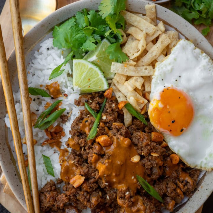 Thai red curry ground beef bowl with a fried egg, peanut sauce, rice, wonton strips, cilantro, and lime wedges