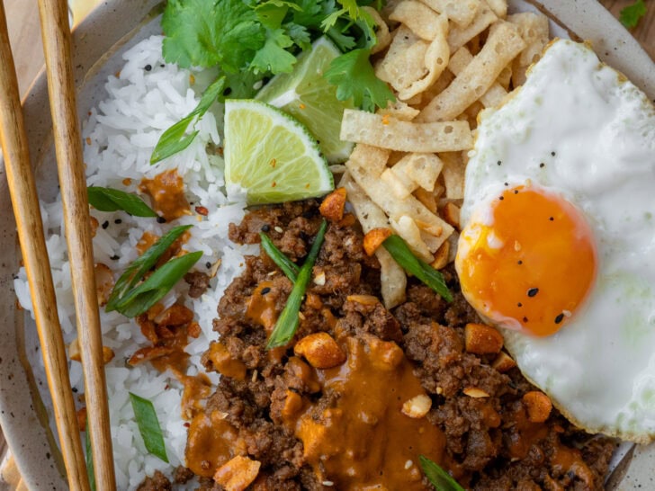 Thai red curry ground beef bowl with a fried egg, peanut sauce, rice, wonton strips, cilantro, and lime wedges