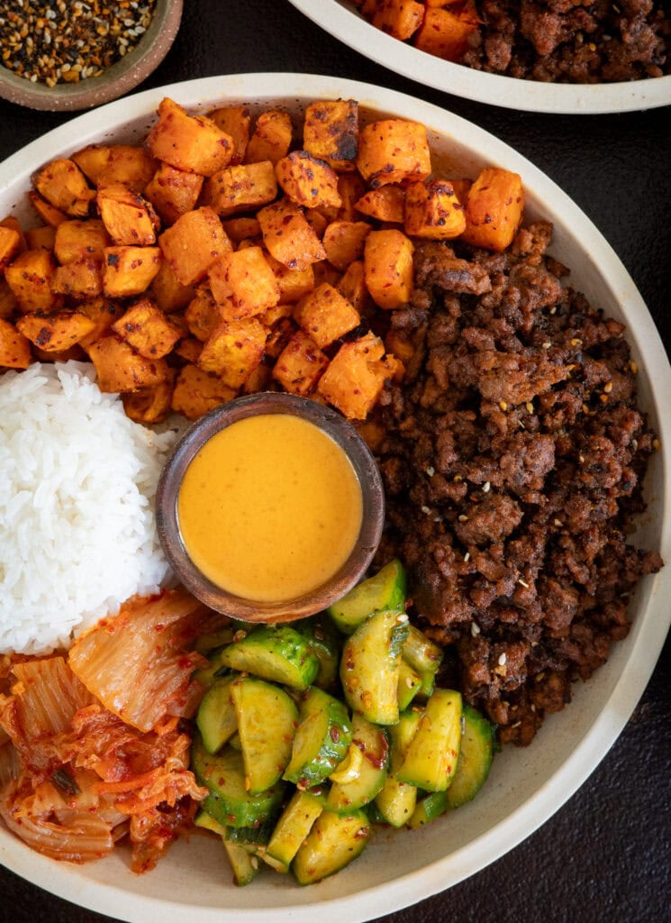 Korean inspired ground beef and sweet potatoes in a bowl with kimchi, pickled cucumbers, rice, and ssamjang mayo