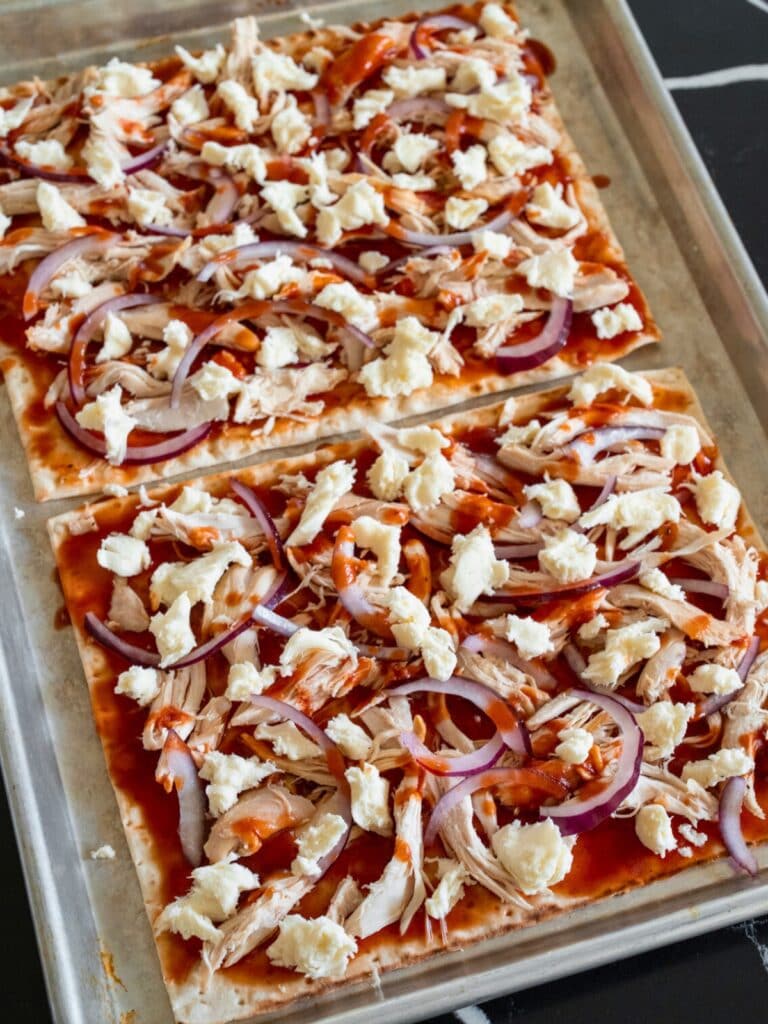 two lavash flatbreads on a half sheet pan topped with bbq sauce, pulled rotisserie chicken, sliced red onion, and fresh mozzarella