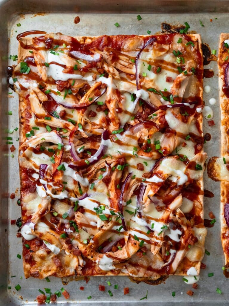 baked chicken flatbread on a sheet pan garnished with crispy bacon, chives, and a drizzle of ranch and bbq sauce