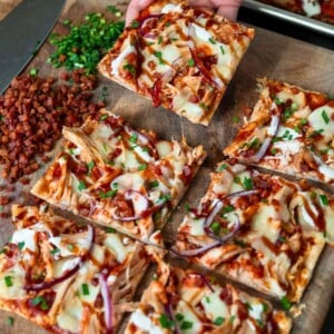 picking up a piece of a chicken flatbread off a cutting board with chopped bacon and chives