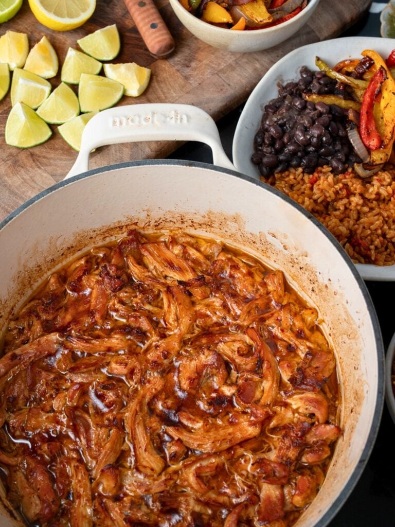pulled Mexican chicken in a Dutch oven next to burrito bowl with fajita veggies and sliced lemons and limes on a cutting board