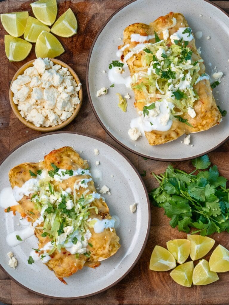 two plates with green chile rotisserie chicken enchiladas garnished with Greek yogurt, shredded lettuce, cilantro, and queso fresco
