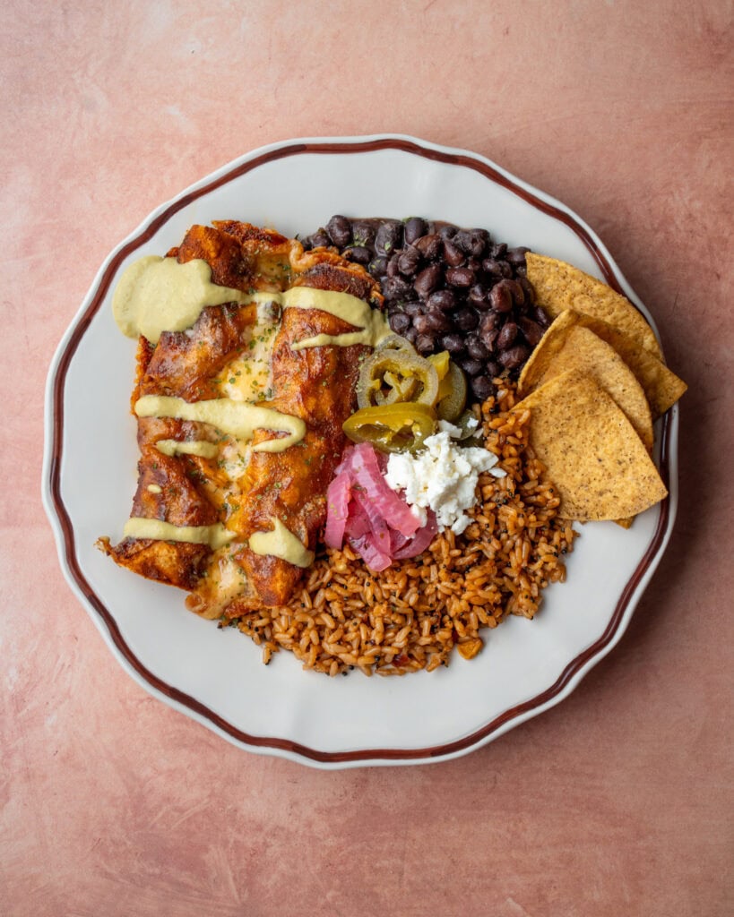 two beefy cottage cheese enchiladas on a plate with rice, beans, tortilla chips, queso fresco, pickled onions, and pickled jalapeno peppers