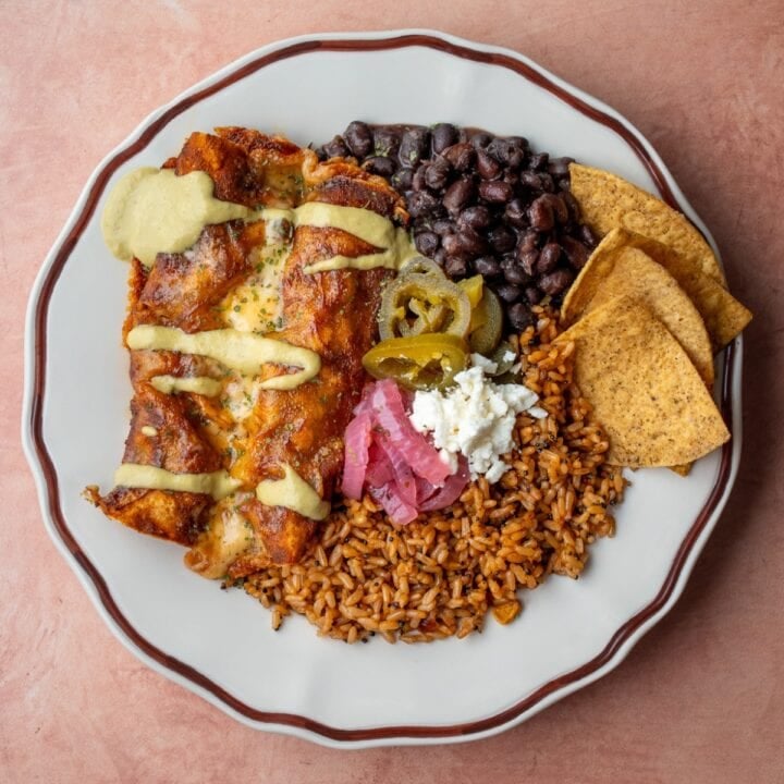 two beefy cottage cheese enchiladas on a plate with rice, beans, tortilla chips, queso fresco, pickled onions, and pickled jalapeno peppers