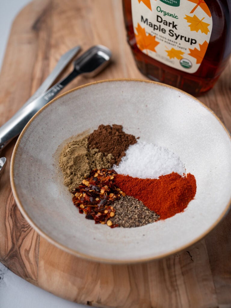 maple syrup bottle next to a bowl with sage, allspice, smoked paprika, kosher salt, black pepper, and red pepper flakes