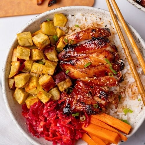 sliced air fried chicken thigh in a bowl with steamed rice, roasted japanese sweet potatoes, pickled cabbage and carrots, and toasted sesame seeds